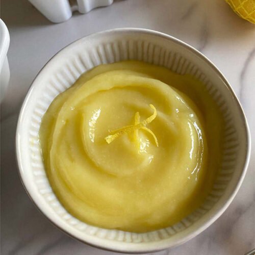 A bowl of citrusy lemon curd topped with zest on a marble countertop