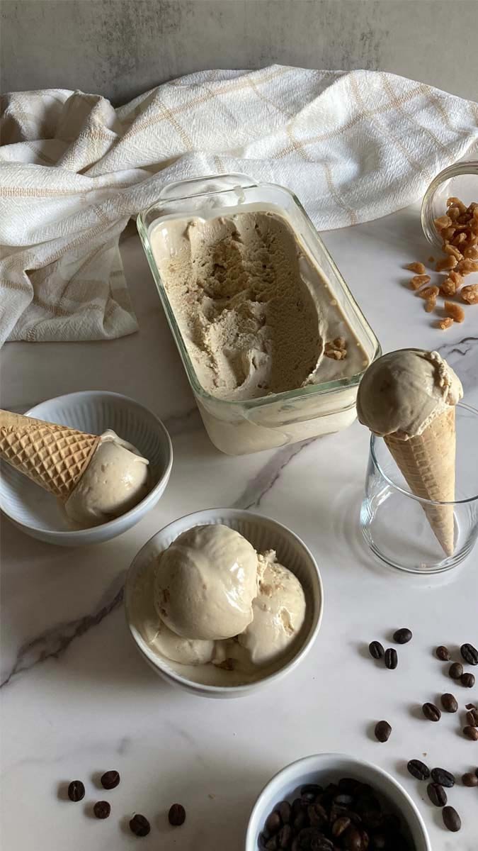 several creamy scoops of coffee toffee ice cream in cones, a bowl, and container with scattered coffee beans and toffee pieces