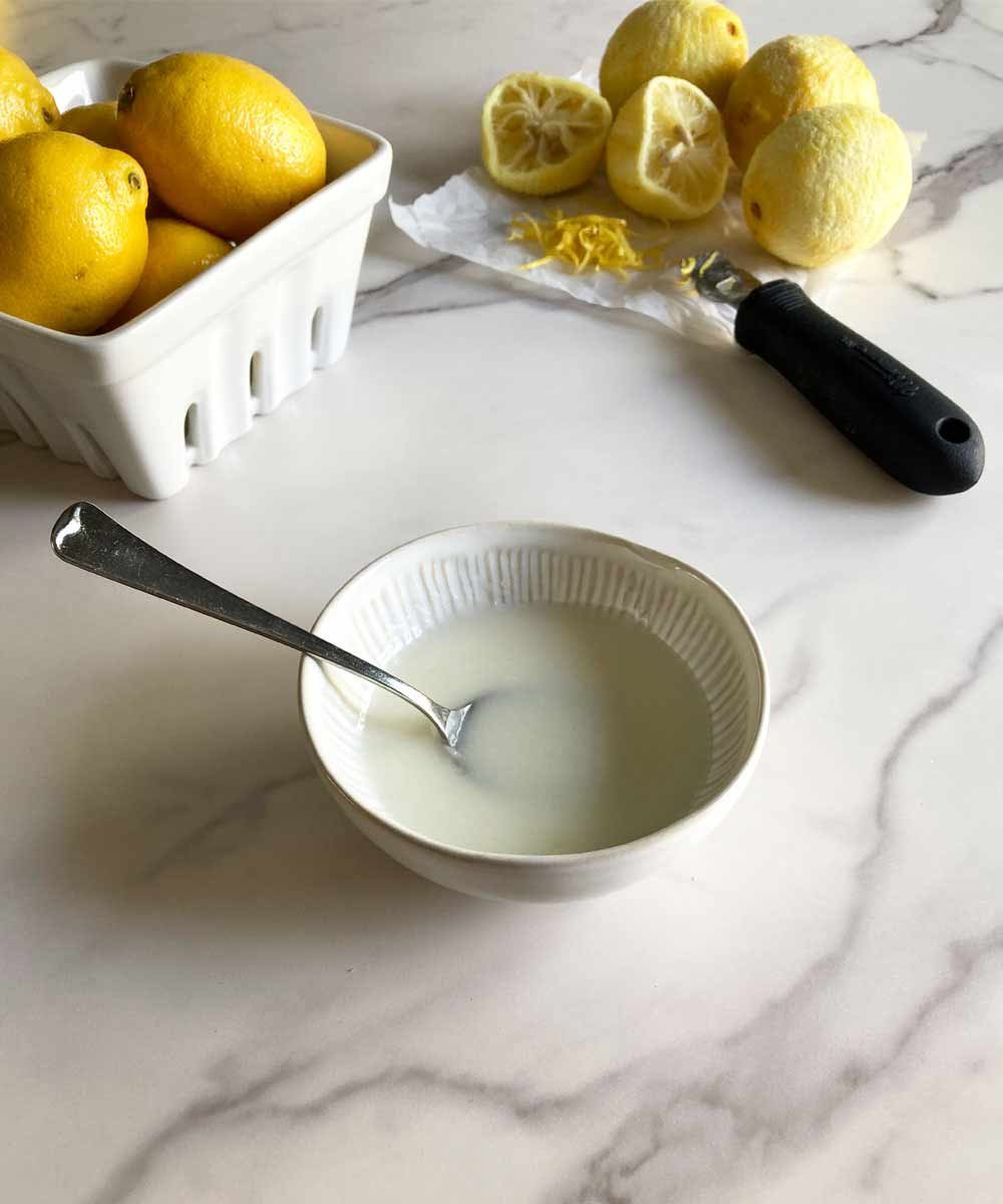 A bowl of lemon glaze surrounded by whole and zested lemons on a marble counter