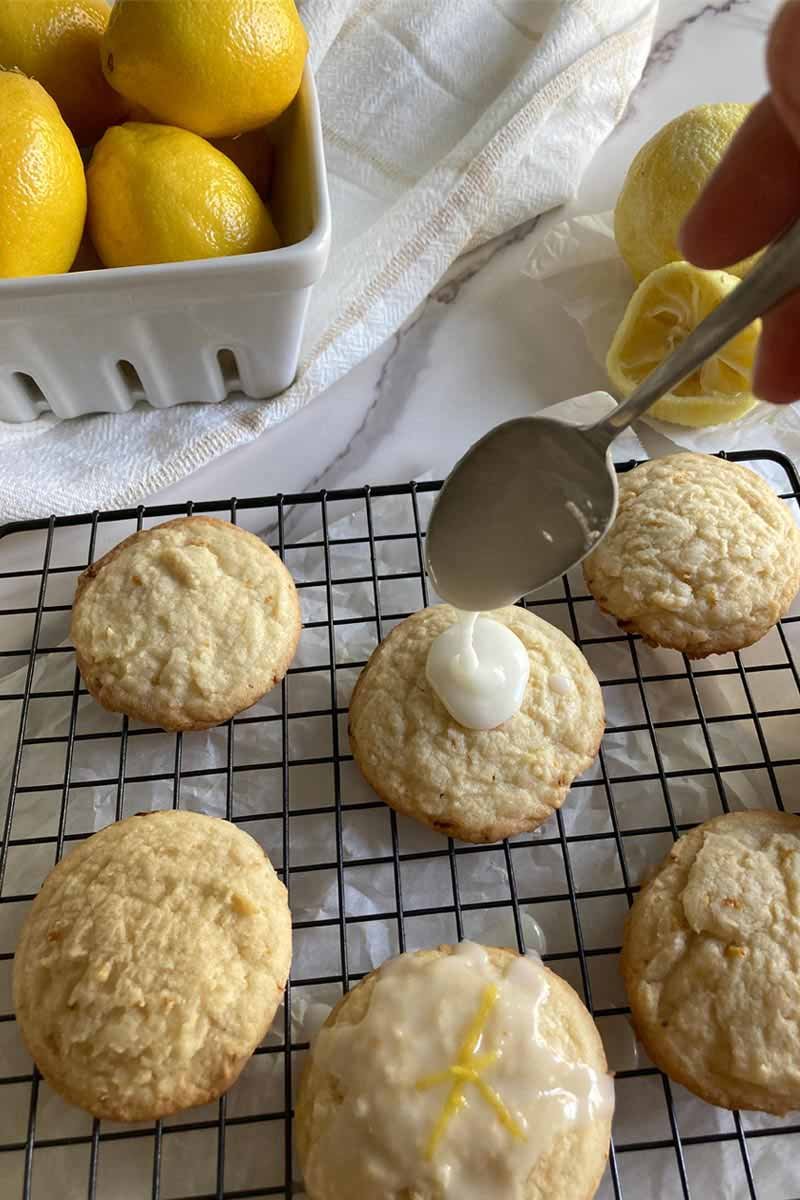 A cooling rack filled with lemon cookies, one has lemon glaze being poured on top