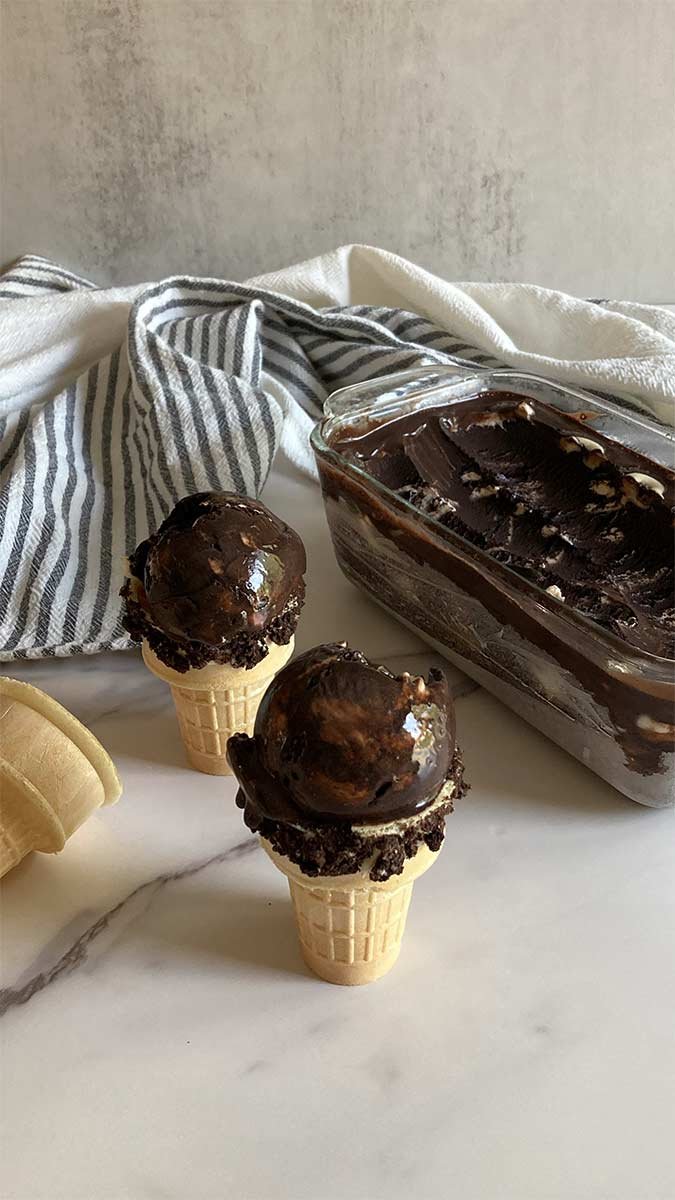 two scoops of creamy black cocoa cookies and cream ice cream in sugar cones, that have been dipped in Oreo cookie crumbs, sitting on a marble countertop with a container of ice cream and a striped towel behind it