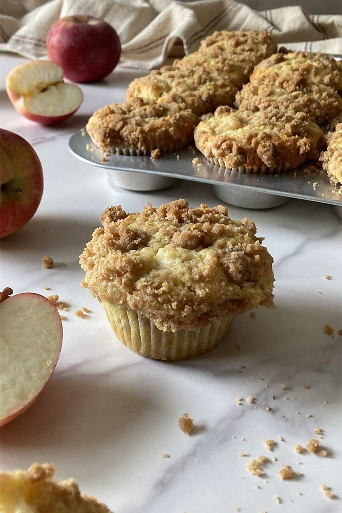 single moist apple muffin with cinnamon streusel topping sitting on a counter with a pan of muffins in the background