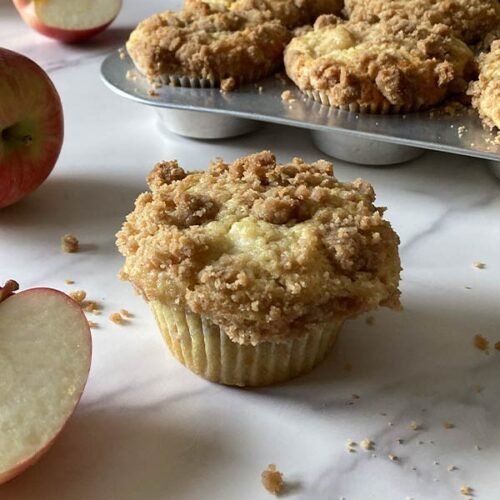 moist apple muffin with cinnamon streusel topping sitting on a counter with a pan of muffins in the background