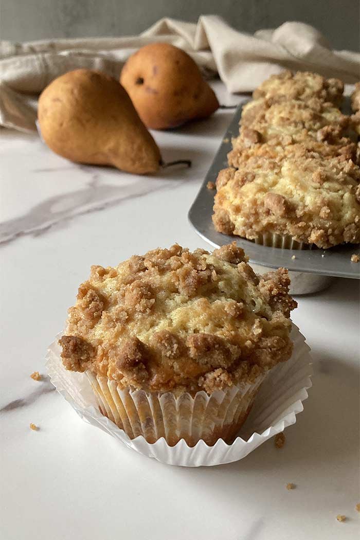 moist pear streusel muffin in a cupcake wrapper sitting on a marble counter with a muffin pan and pears