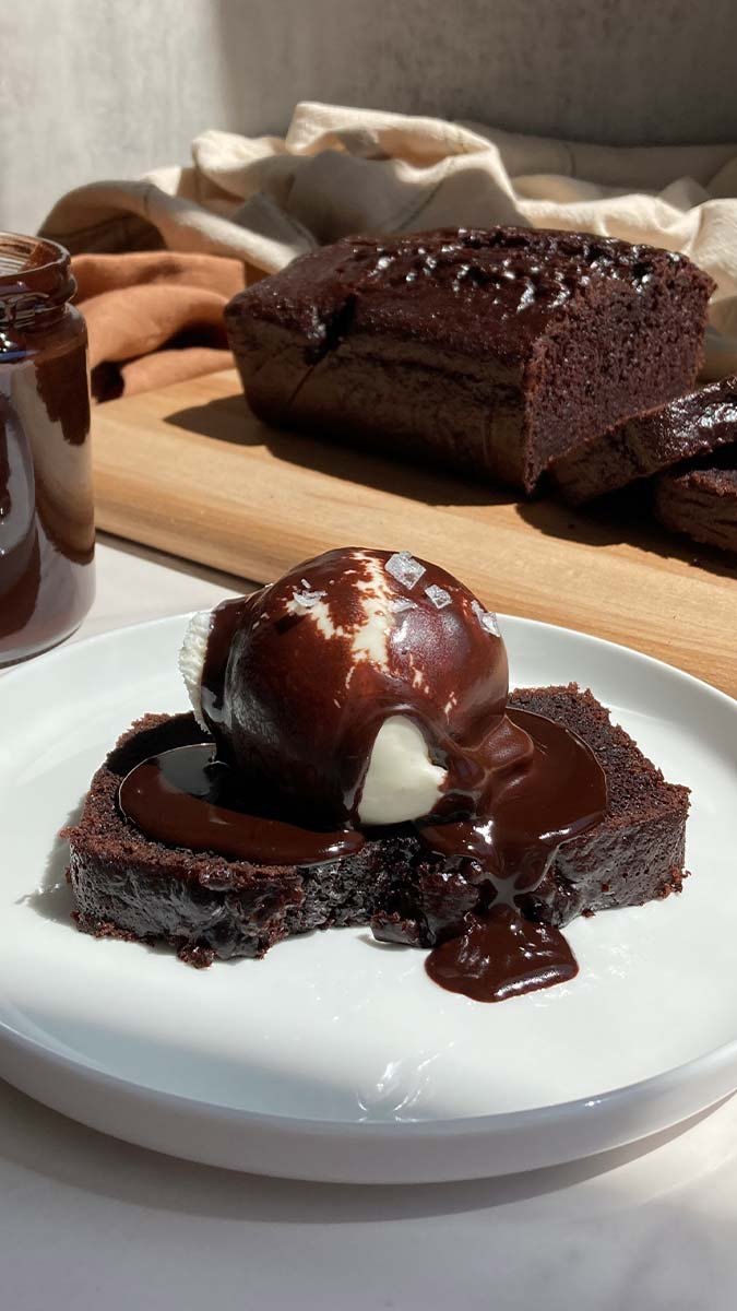 slice of chocolate espresso loaf cake with a scoop of vanilla ice cream, brown butter espresso hot fudge, and flaky salt with the sliced loaf in the background