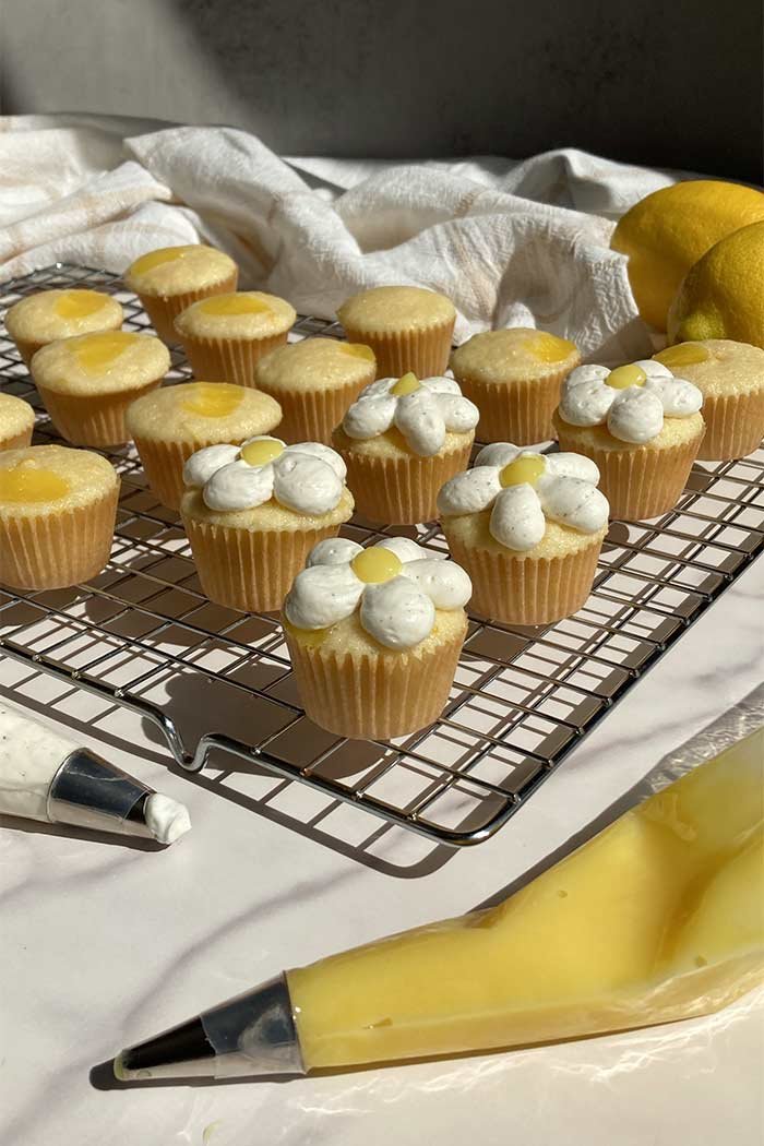 mini lemon chantilly cupcakes on a cooling rack surrounded by piping bags filled with whipped cream frosting and lemon curd