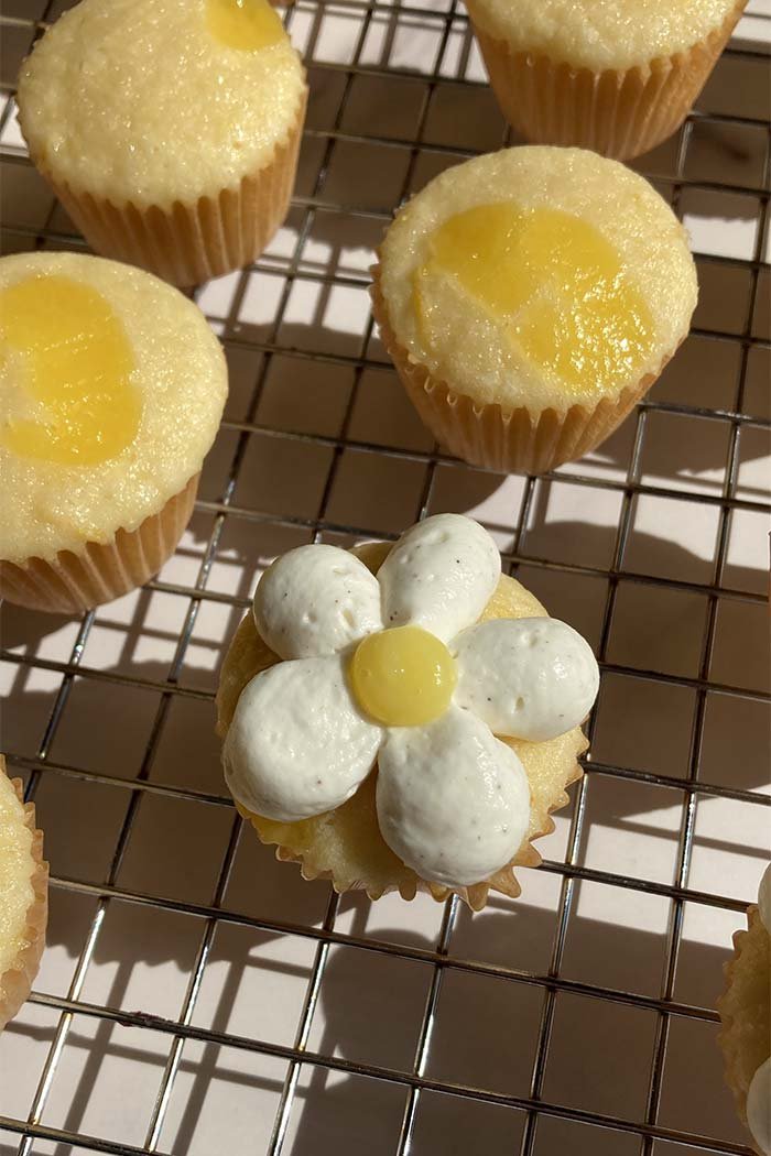 a frosted lemon chantilly cupcake on a cooling rack surrounded by unfrosted cupcakes topped with lemon curd