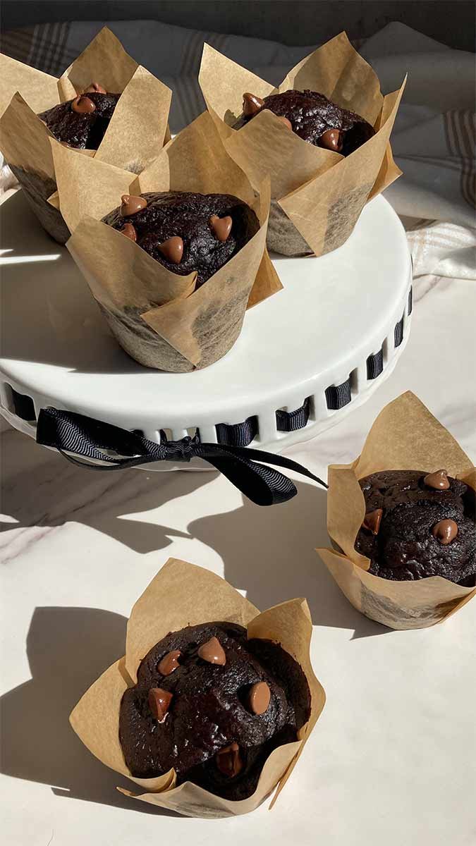 two homemade triple chocolate muffins on a countertop next to a cake stand filled with more muffins