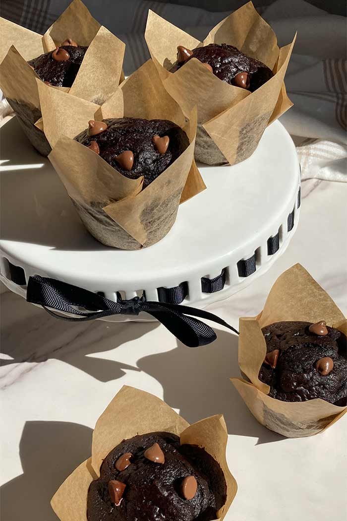 two homemade triple chocolate muffins on a countertop next to a cake stand filled with more muffins