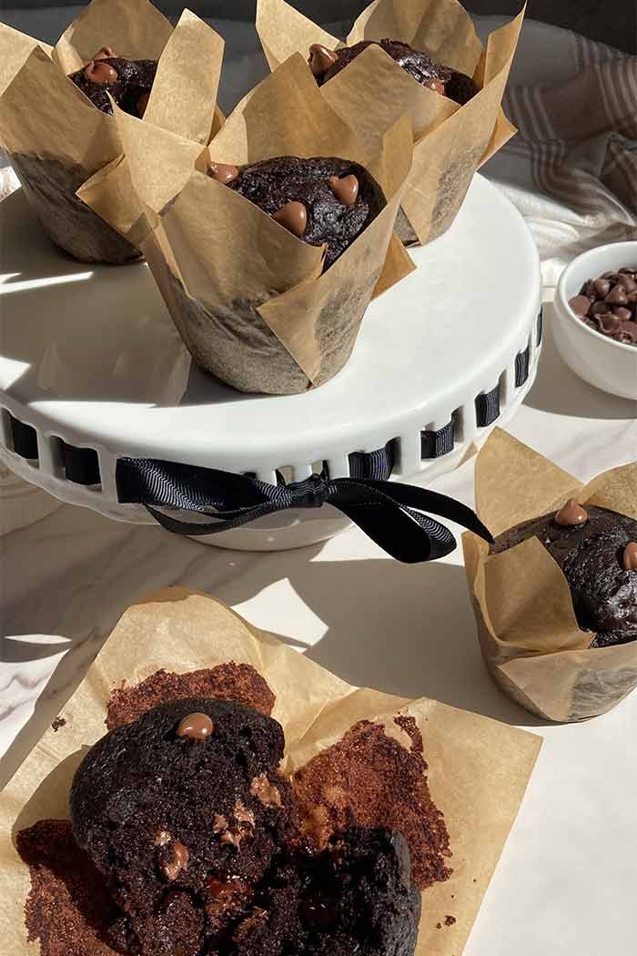 homemade triple chocolate muffins on a countertop, one broken in half to show the moist crumb, next to a cake stand filled with more muffins
