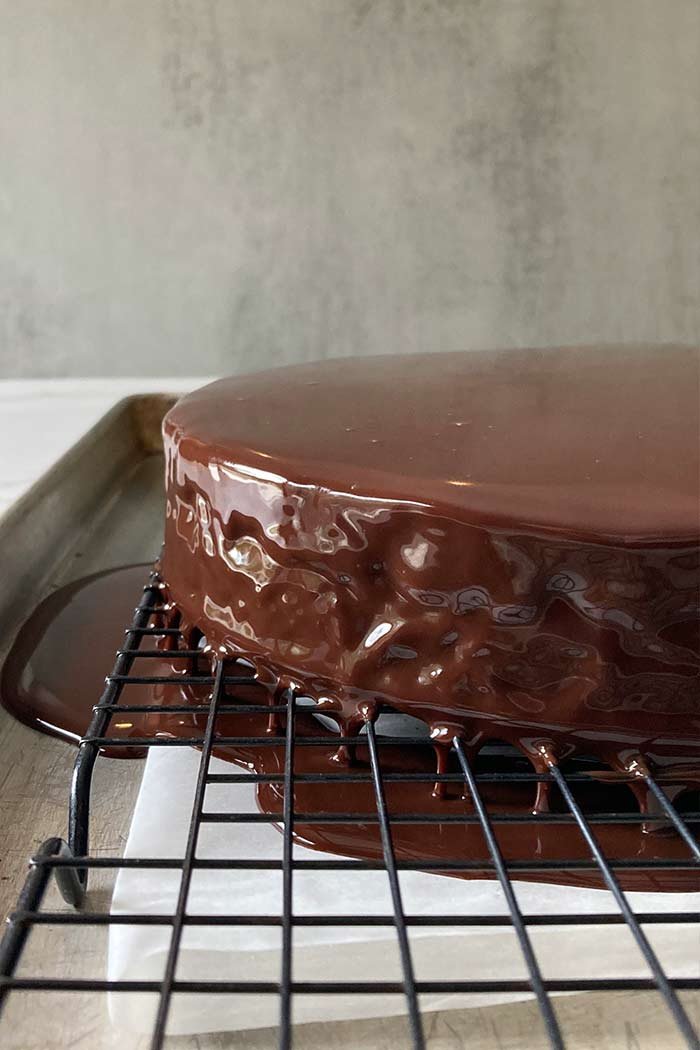 fresh chocolate ganache poured over a peanut butter mousse brownie crunch cake on a wire rack